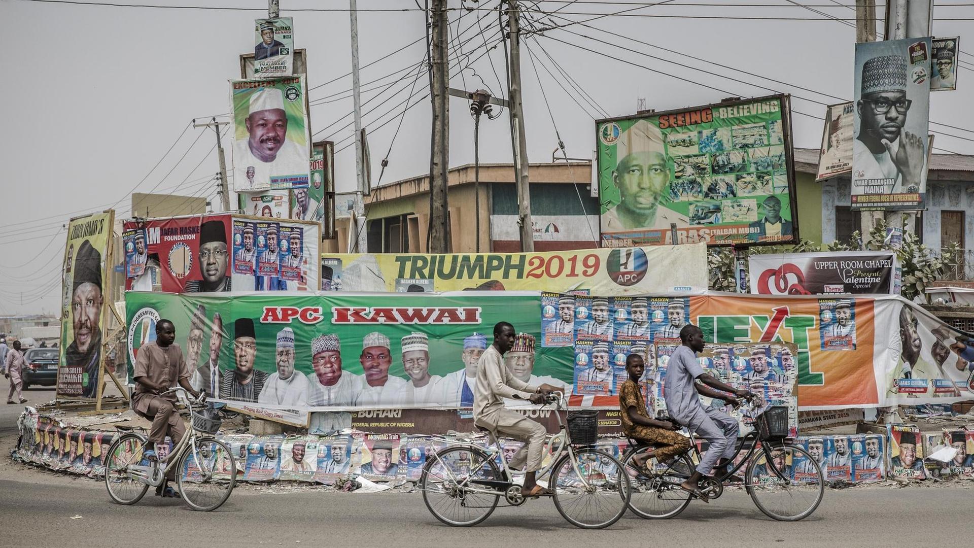 Menschen fahren auf Räder in der nigerianischen Stadt Mubi im Staat Adamawa an Wahlkampf-Plakaten vorbei. Die Wahl wird sich vor allem zwischen Präsident Muhammadu Buhari und seinem wichtigsten Herausforderer, Atiku Abubakar, entscheiden.
