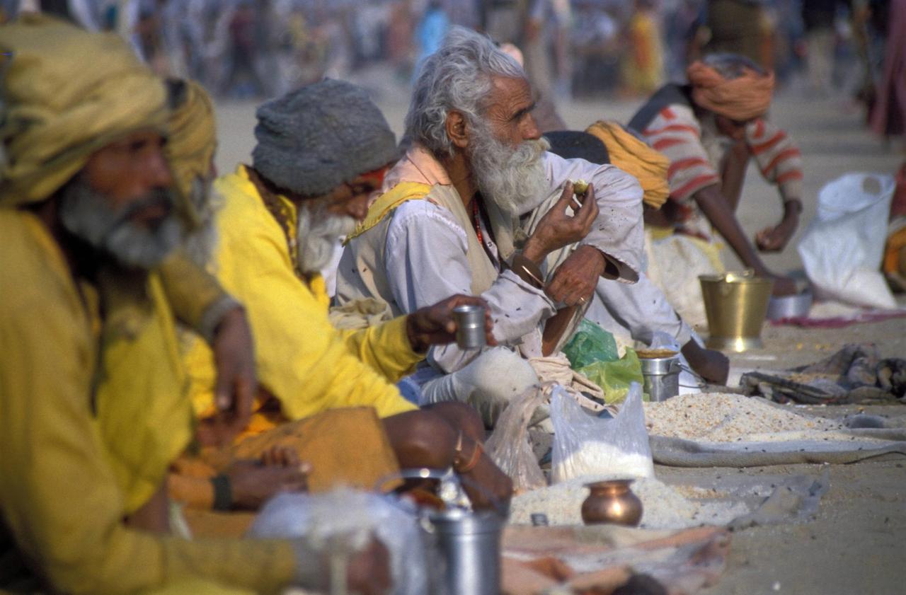 Hinduistische Bettelmönche in langen Gewändern sitzen auf dem Boden. Vor ihnen liegen ausgebreitete Tücher mit essen.