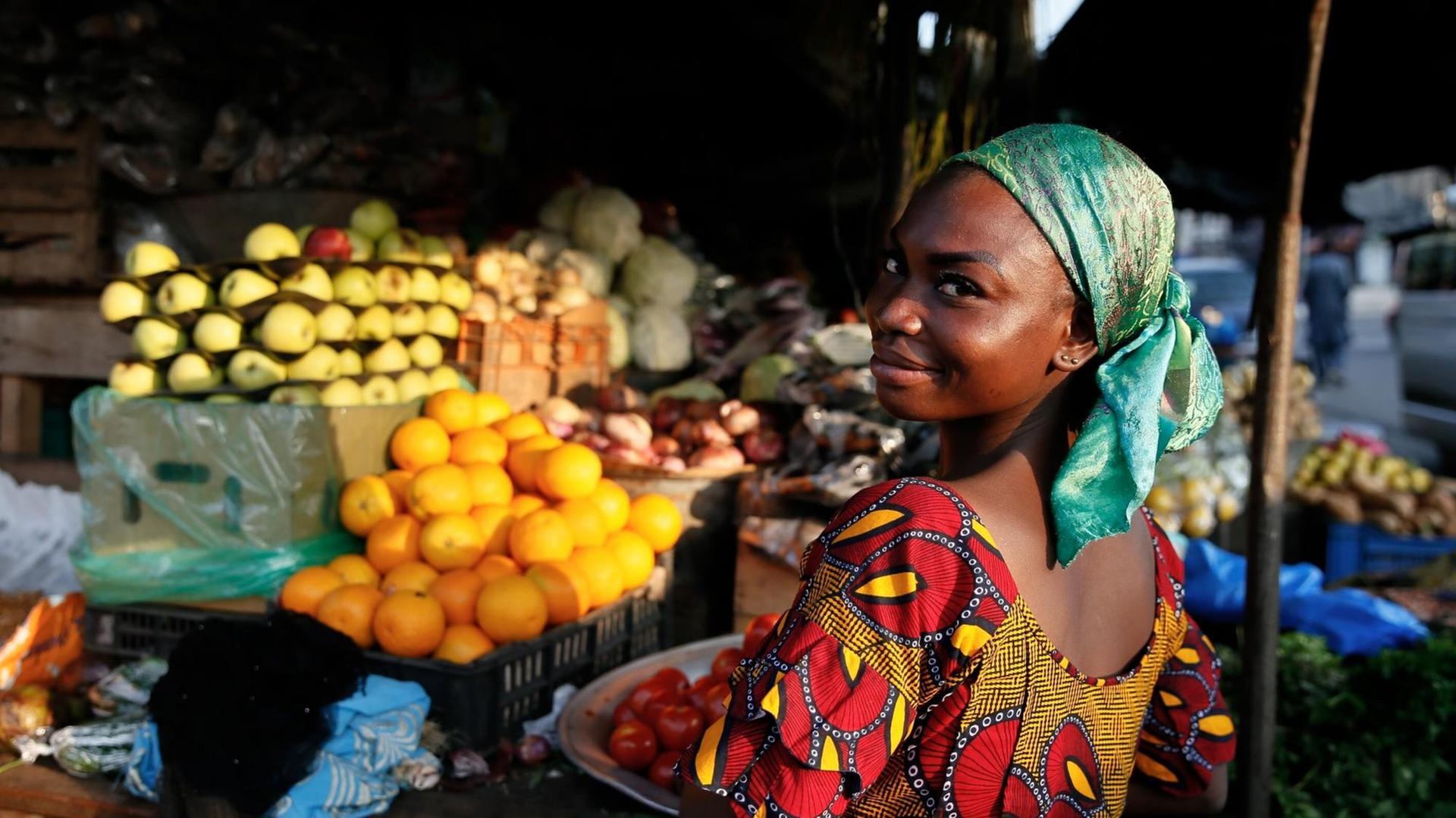 Eine Obstverkäuferin blickt über die Schulter in die Kamera. Im Hintergrund sind Obststände mit reifen Früchten zu sehen.