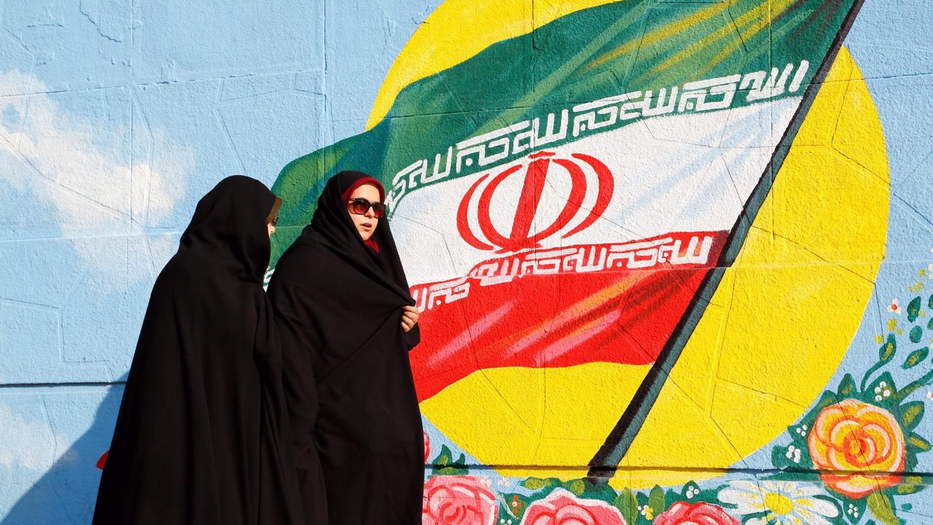 Zwei iranische Frauen in Teheran vor einer iranischen Flagge, die als Graffiti an eine Wand gesprüht ist Zwei iranische Frauen in Teheran vor einer iranischen Flagge, die als Graffiti an eine Wand gesprüht ist