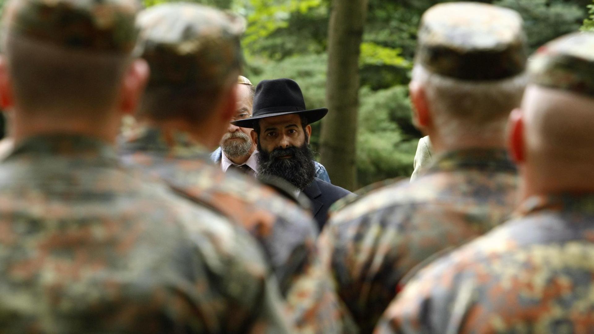 Man sieht die Soldaten in Tarnuniformen von hinten. Zwischen ihren Schultern sieht man einen jüdischen Mann mit dunklem Vollbart und Hut. Im Hintergrund Bäume. 