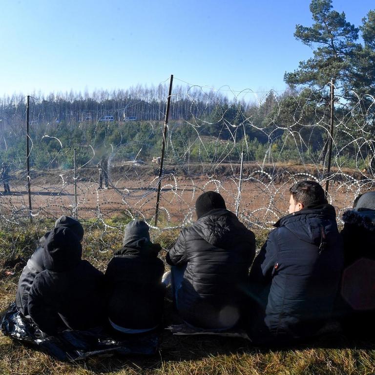 Fünf Personen sitzen an der belaruissisch-polnischen Grenze vor einem Stacheldrahtzaun