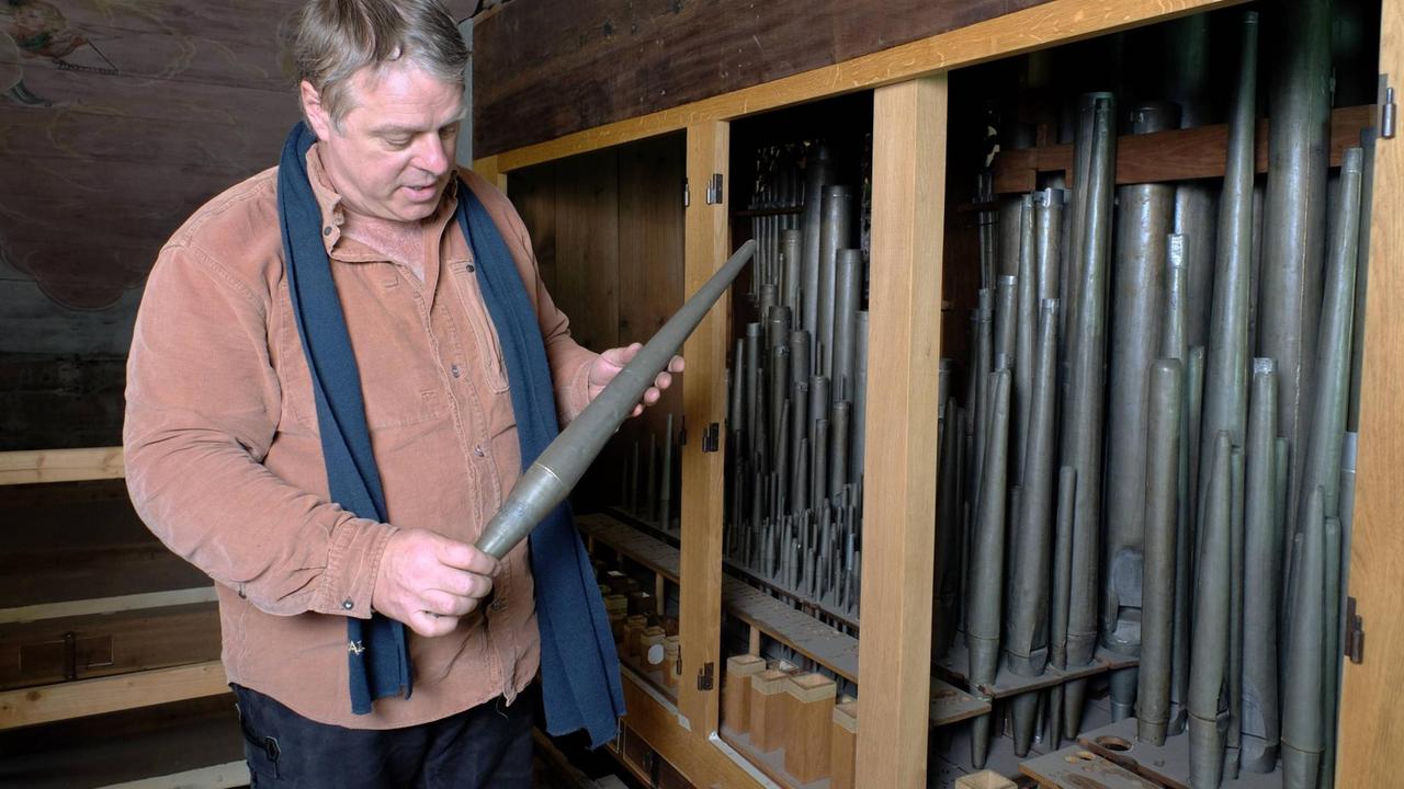 Ein Mann steht neben einem offenen Orgelkasten, in dem verschieden große, verstaubte Orgelpfeifen stehen. Er hat eine der alten Zinnpfeifen in der Hand und betrachtet ihren Zustand.