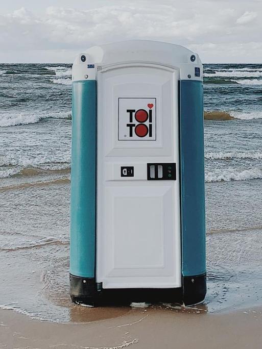 Ein transportables Toilettenhäuschen steht in der Meeresbrandung am Strand.