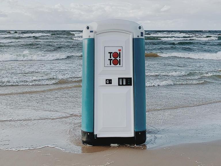Ein transportables Toilettenhäuschen steht in der Meeresbrandung am Strand.