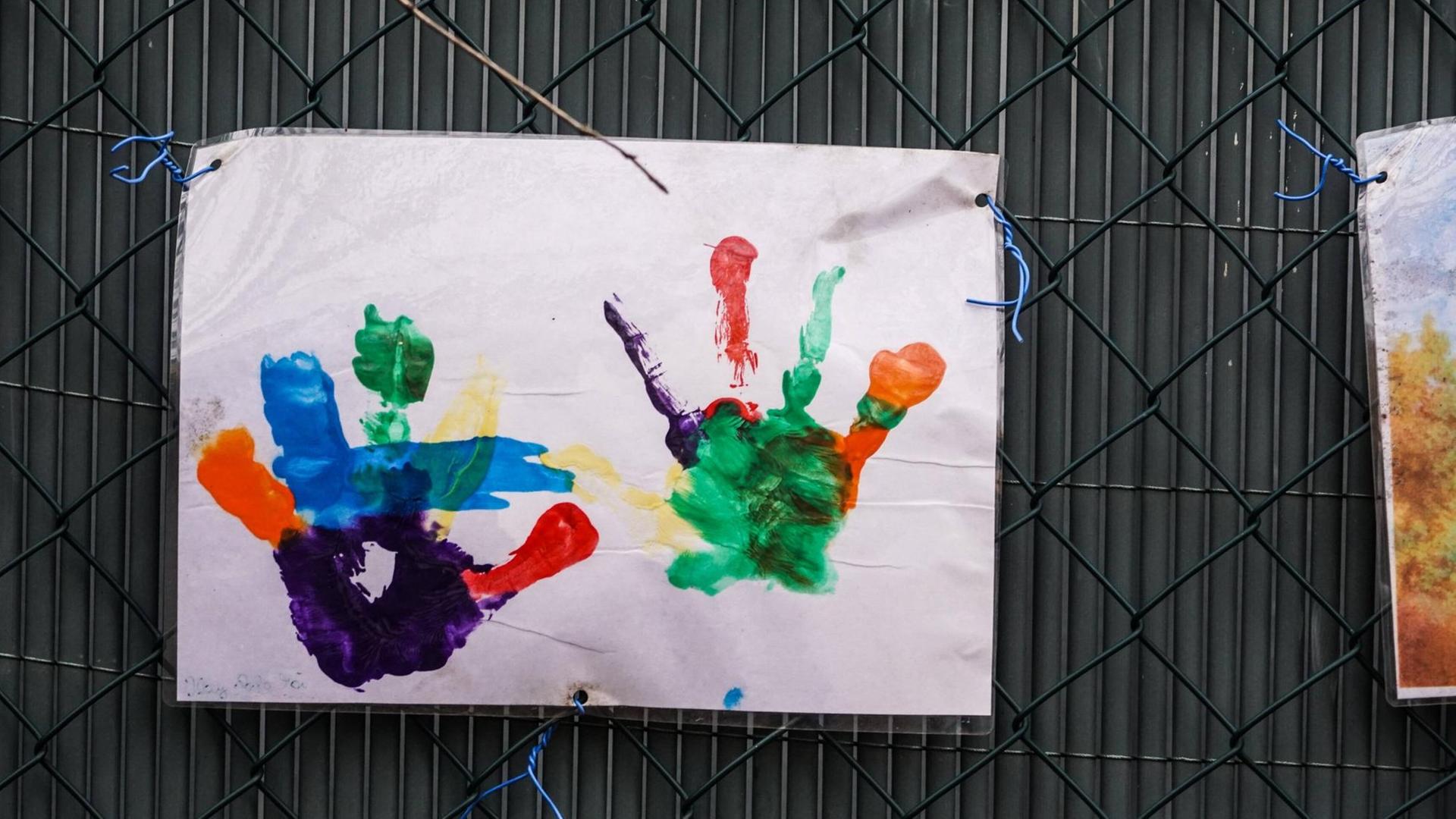 An einem Zaun hängt ein Blatt Papier mit dem vielfarbigen Abdruck zweier Kinderhände.