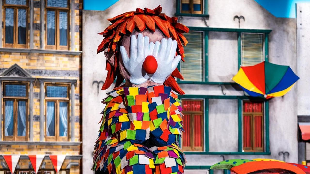 Eine Clowns-Figur sitzt mit den Händen vor dem Gesicht auf einem Rosenmontagswagen, links unten ist ein Modell eines Coronavirus zu sehen, im Hintergrund Modelle von Häusern