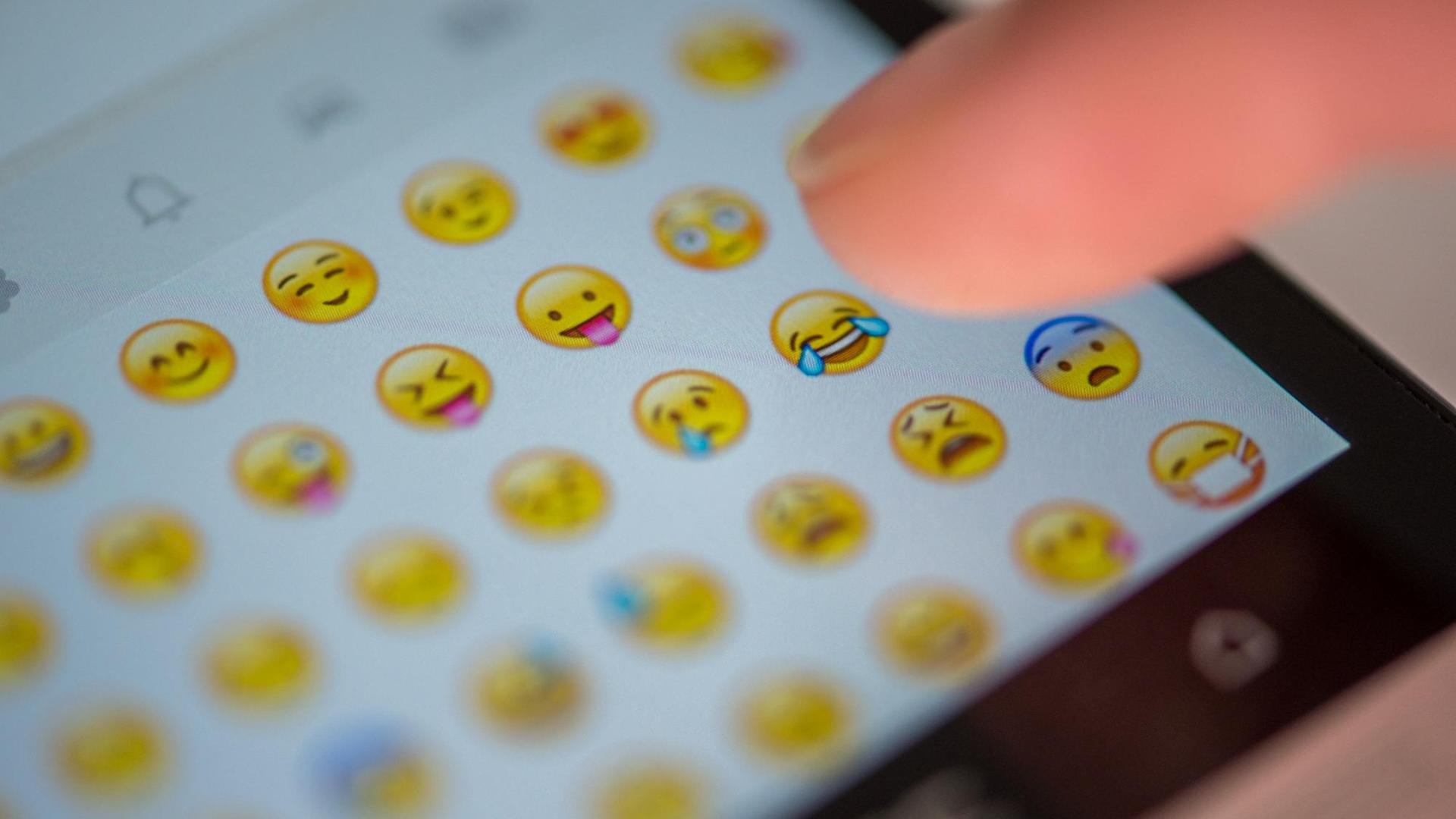 Eine Frau tippt auf den Bild-Schirm von einem Handy. Man kann viele Smiley-Emojis sehen.