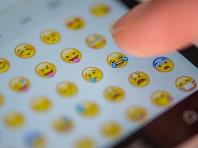 Eine Frau tippt auf das Display eines Smartphones, auf dem zahlreiche Emojis zu sehen sind