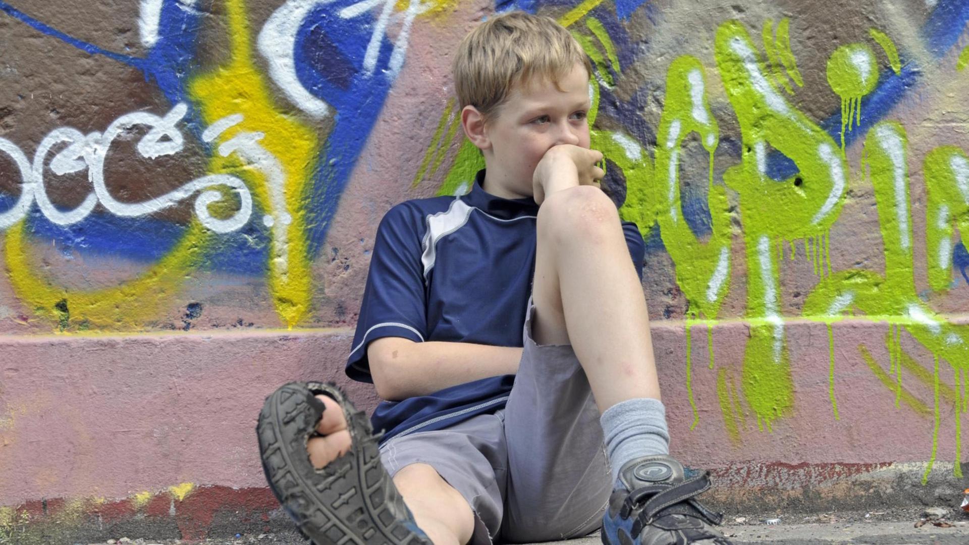 Ein Junge mit kaputten Schuhen sitzt auf dem Boden vor einer bunten Wand.