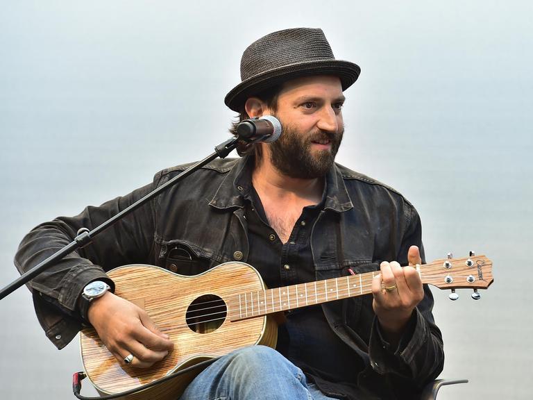 Der Musiker Daniel Kahn sitzt mit schwarzem Hemd, schwarzem Hut und einer Ukulele hinter einem Mikrofon.