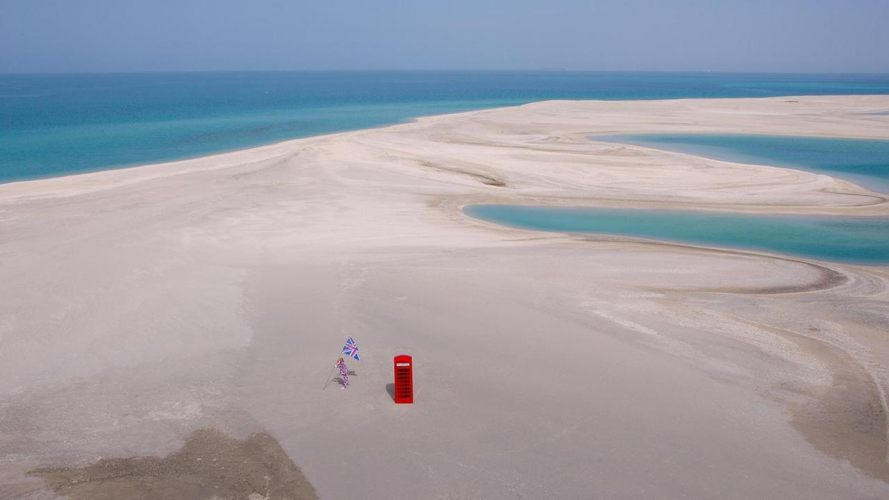 In Vogelperspektive ist eine flache Sandfläche zu erkennen umgeben von türkisfarbenem Wasser. Im Vordergrund steht eine rote, typisch britische Telefonzelle, links daneben ein Mann in Union-Jack Anzug und Flagge.