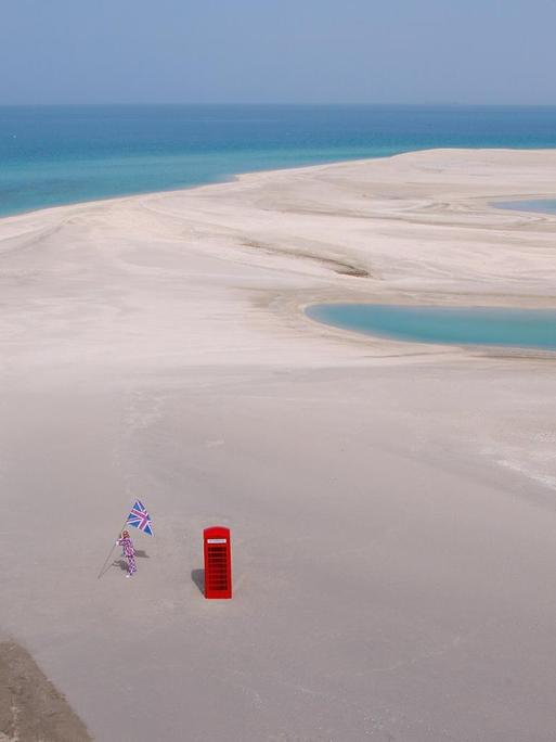 In Vogelperspektive ist eine flache Sandfläche zu erkennen umgeben von türkisfarbenem Wasser. Im Vordergrund steht eine rote, typisch britische Telefonzelle, links daneben ein Mann in Union-Jack Anzug und Flagge.