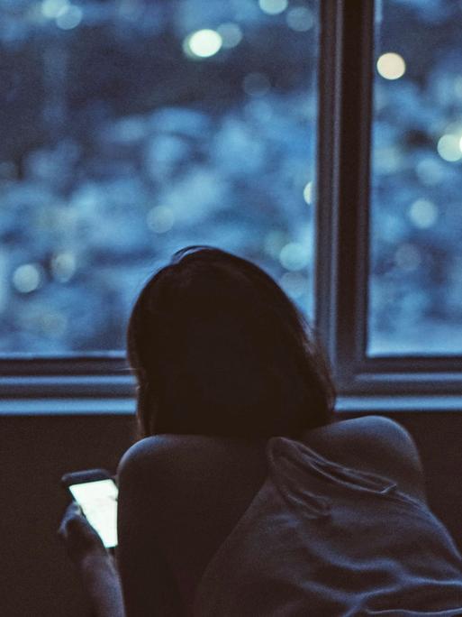 Eine junge Frau liegt bäuchlings im Bett und schaut auf ihr Smartphone.