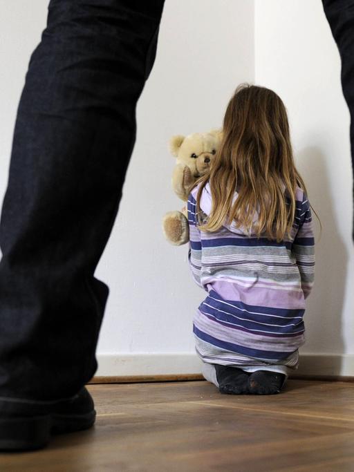 Symbolfoto zum Thema Kindesmissbrauch: Man sieht ein Mädchen in einer Zimmerecke mit Teddy von hinten und im Vordergrund die Beine eines Mannes