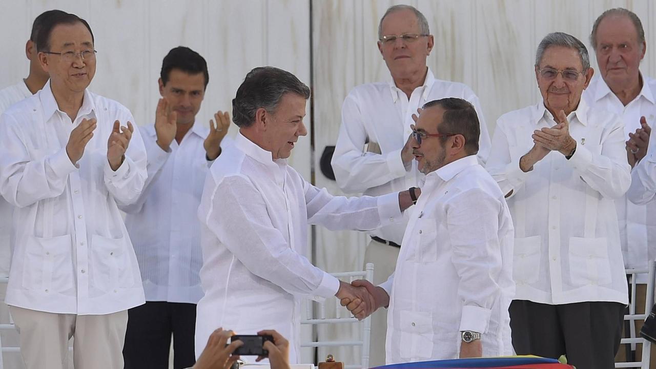 Kolumbiens Präsident Manuel Santos und der Anführer der kolumbianischen Guerilla-Organisation FARC, Timoleón Jiméne besiegeln am 26.09.2016 mit einem Handschlag das Friedensabkommen.
Hinter den beiden Männern, stehen applaudierende Menschen, darunter UNO-Generalsekretär Ban Ki-moon. Alle tragen weiße Hemden. Das Bild enstand bei der Zeremonie zur Unterzeichnung des Friedensvertrages.