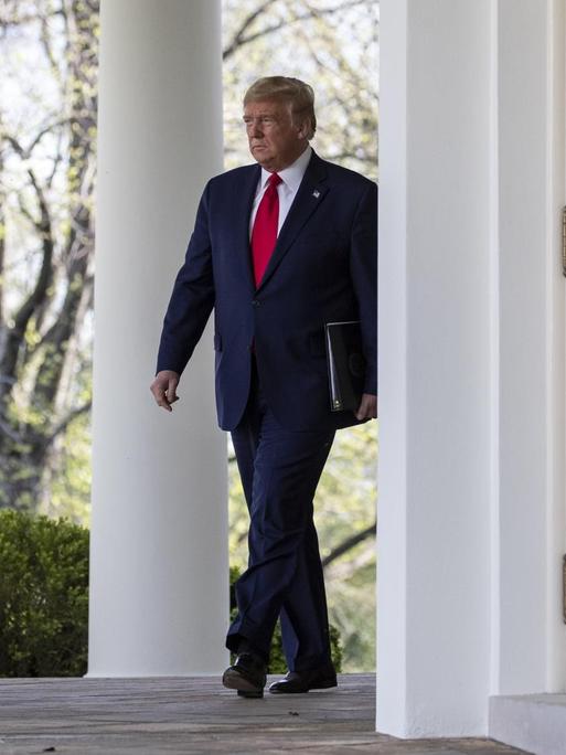 Trump läuft mit einem Ordner im Arm auf einer Terasse des Weißen Hauses an dessen Fassade entlang. Im Vordergrund unscharf die Flaggen der USA und des Präsidenten.