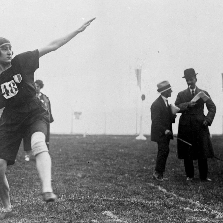 Ein Schwarzweiß-Foto zeigt eine Athletin mit einer Kappe, die mit einem Ausfallschritt Anlauf zum Kugelstoßen nimmt. Im Hintergrund sind zwei Männer mit Borsalino-Hüten ins Gespräch vertieft zu sehen