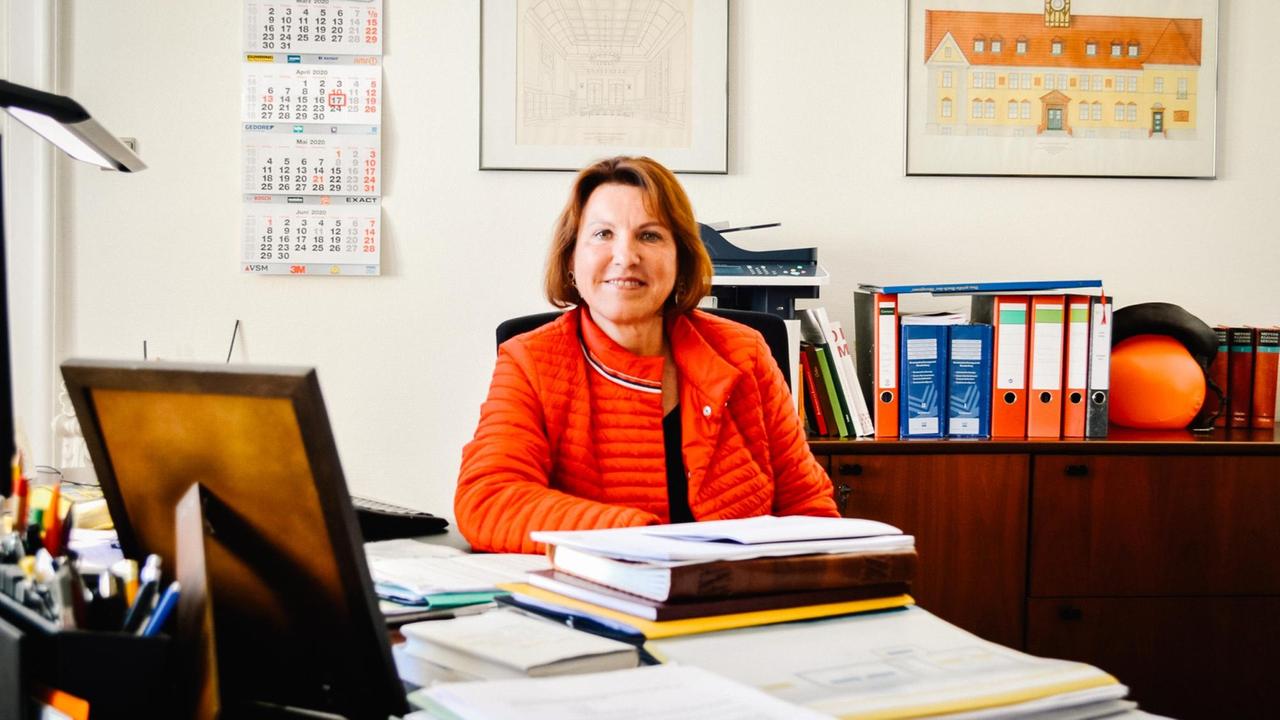 Ines Hübner (SPD), Bürgermeisterin von Velten, sitzt an einem Schreibtisch. Im Vordergrund die Rückansicht eines Bilderrahmens, im Hintergrund Aktenordner und ein Bild des Rathauses der Stadt.
