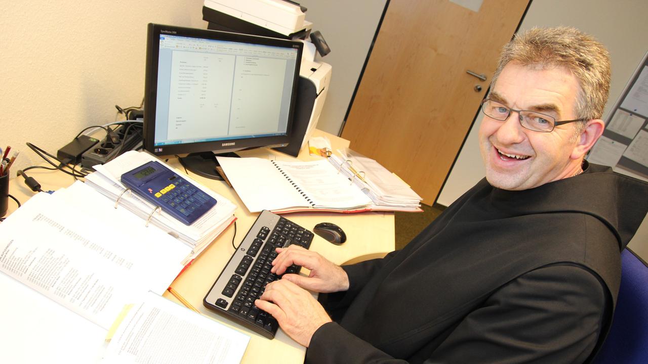 Zu sehen ist Bruder Stephan, der mit einer Kutte bekleidet an seinem Schreibtisch sitzt und am Computer arbeitet. Er lächelt in die Kamera.