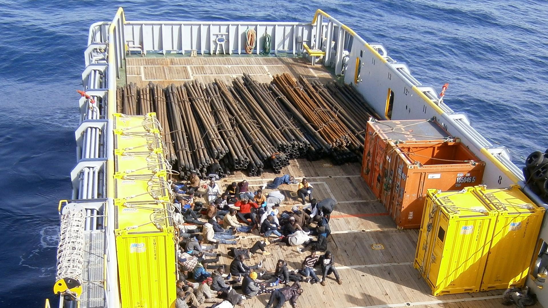 Im hinteren Teil eines Frachtschiffes befinden sich Dutzende Flüchtlinge, hinter ihnen Stahlträger und einige kleinere Container.