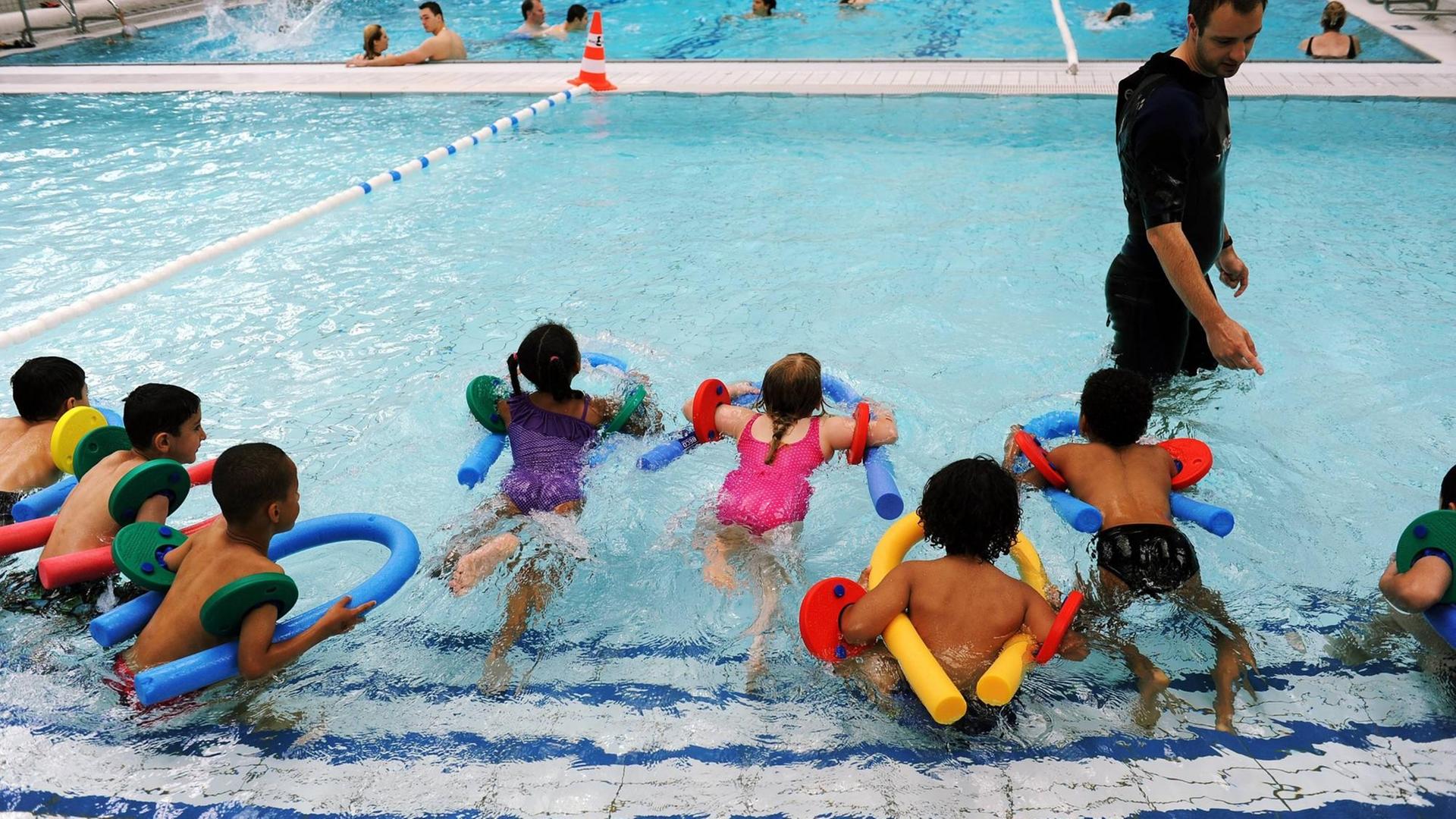 In einem Schwimmbecken stoßen sich knapp zehn Kinder vom Beckenrand im Nichtschwimmerbereich ab. Sie haben Schwimmnudeln. Ein Schwimmlehrer steht im Wasser in schwarzem Short und Shirt vor ihnen.