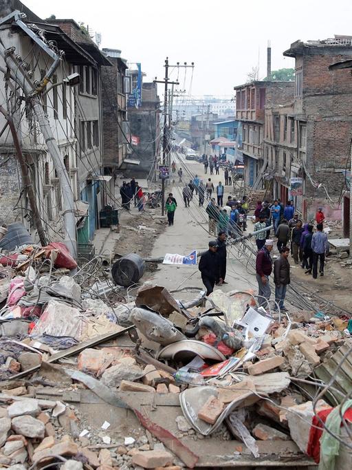 Trümmer eines eingestürzten Hauses in Kathmandu, im Hintergrund der Blick in eine Straße mit zerstörten Häusern und einigen Menschen.