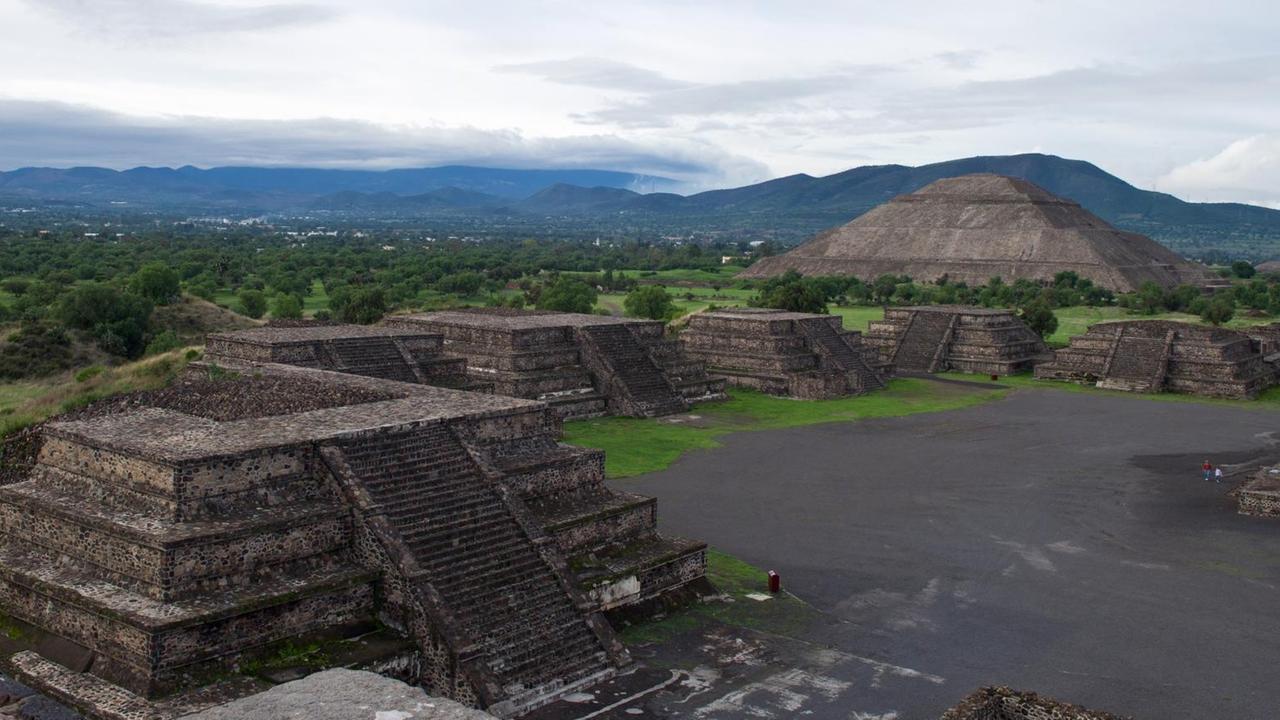 Zwei pyramidenförmige Bauten aus grauem Stein stehen auf einer Fläche. Im Hintergrund sind sehr weit entfernt Berge zu erkennen.