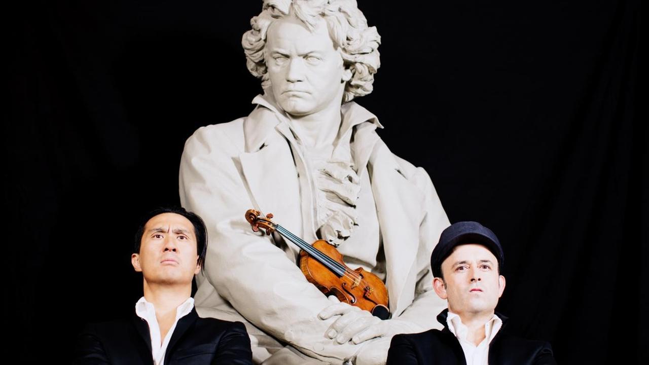 Zwei Männer stehen vor einer Beethovenstatue mit verschränkten Armen und ernsten Blicken. Beethoven hält eine Geige im Arm. Die beiden Männer sind das Comedy-Duo Igudesman und Joo.