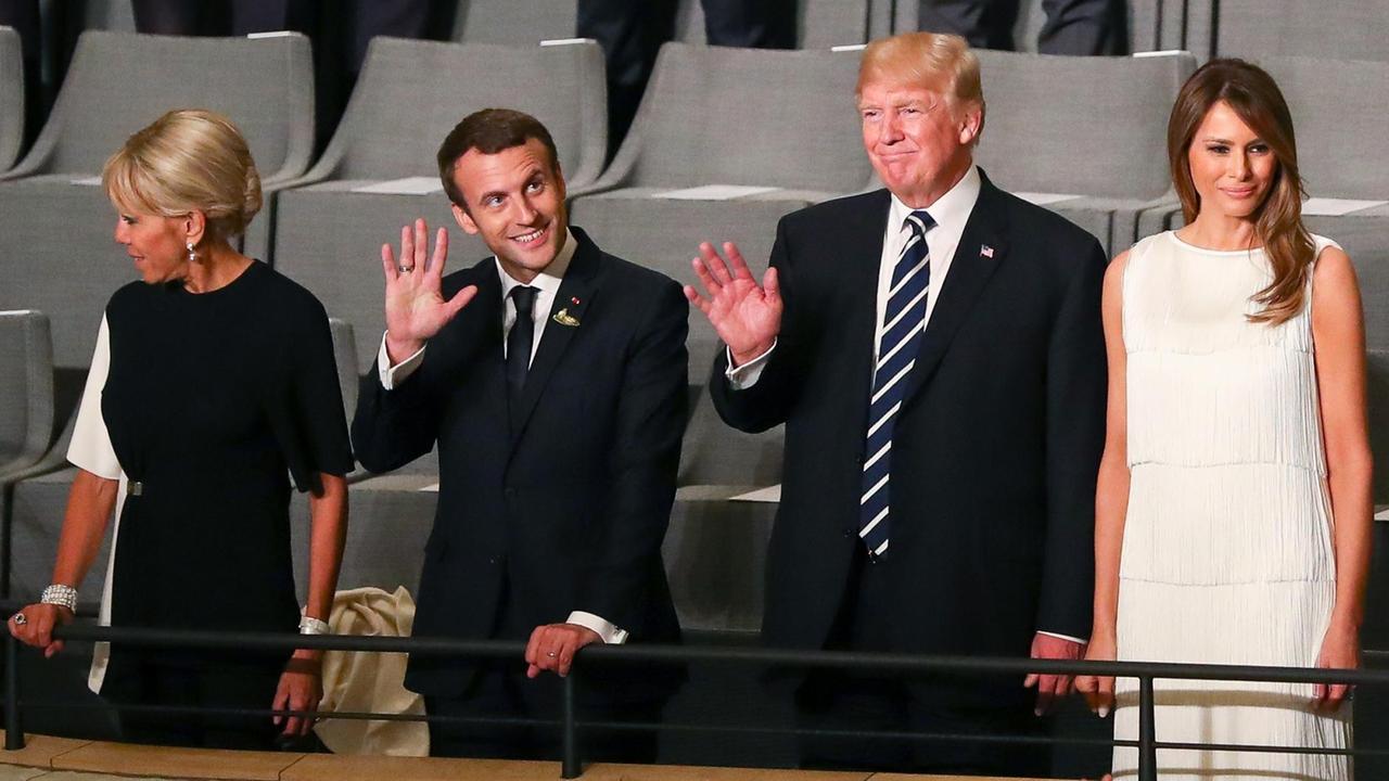 Die vier stehen nebeneinander vor ihren Plätzen an einem Geländer: v. l.n.r. Frau und Herr Macron, Herr und Frau Trump. Die beiden Präsidenten winken lächelnd.