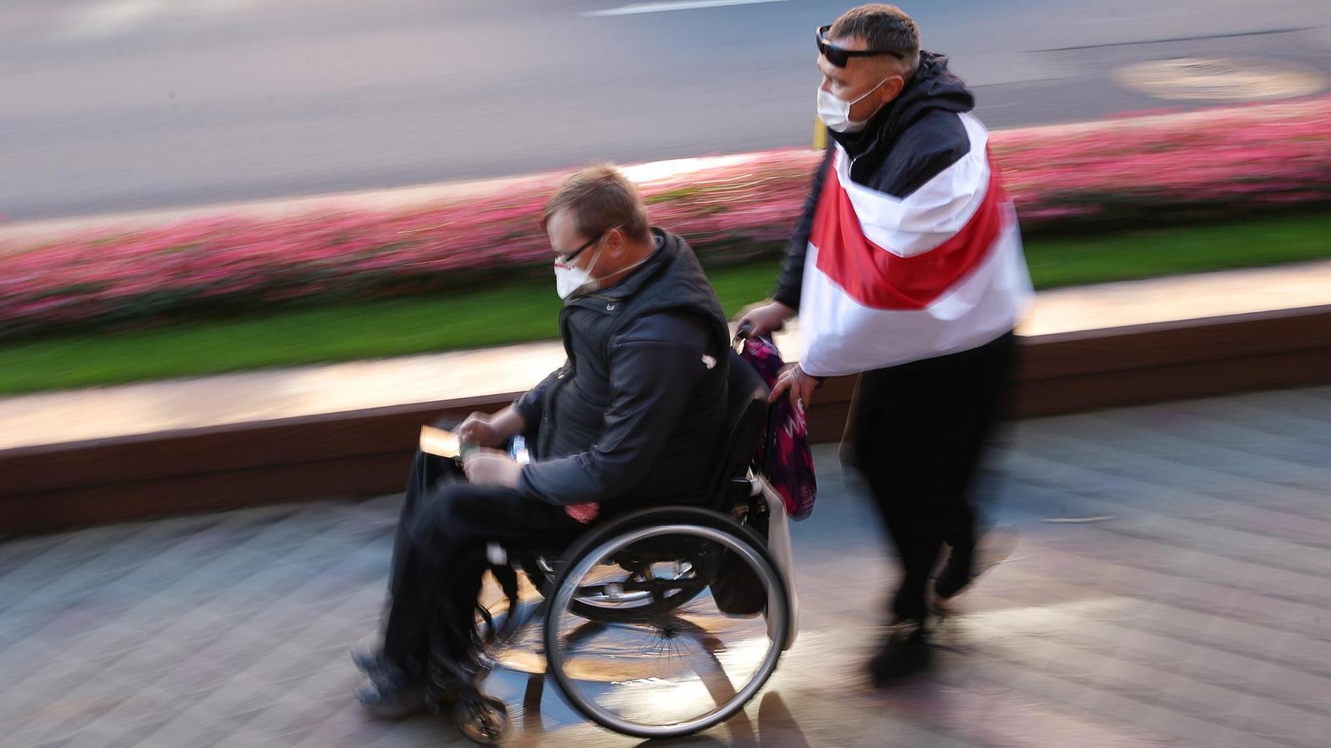 Ein Mann im Rollstuhl wird von einem anderen Mann geschoben, der sich eine weiß-rote Flagge um die Schultern geworfen hat.