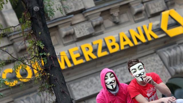 Zwei Demonstranten, die Guy-Fawkes-Masken tragen, sitzen vor dem Logo der Commerzbank.