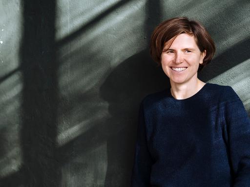 Judith Schalansky steht vor einer Wand, sie schaut freundlich rechts an der Kamera vorbei