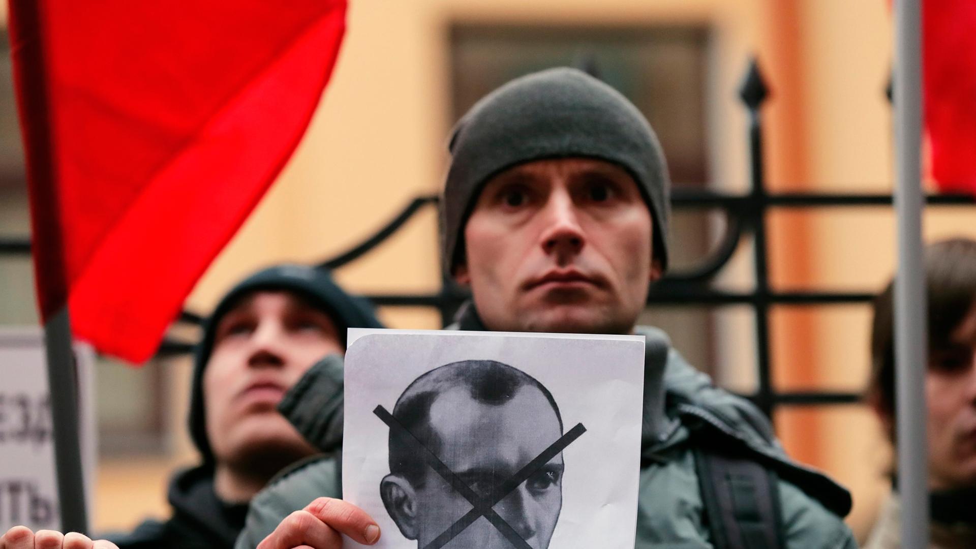 Ein Demonstrant hält ein Schwarz-weiß-Bild von Stepan Bandera in der Hand, dessen Gesicht darauf durchgestrichen ist, im Hintergrund weitere Demonstranten und rote Fahnen