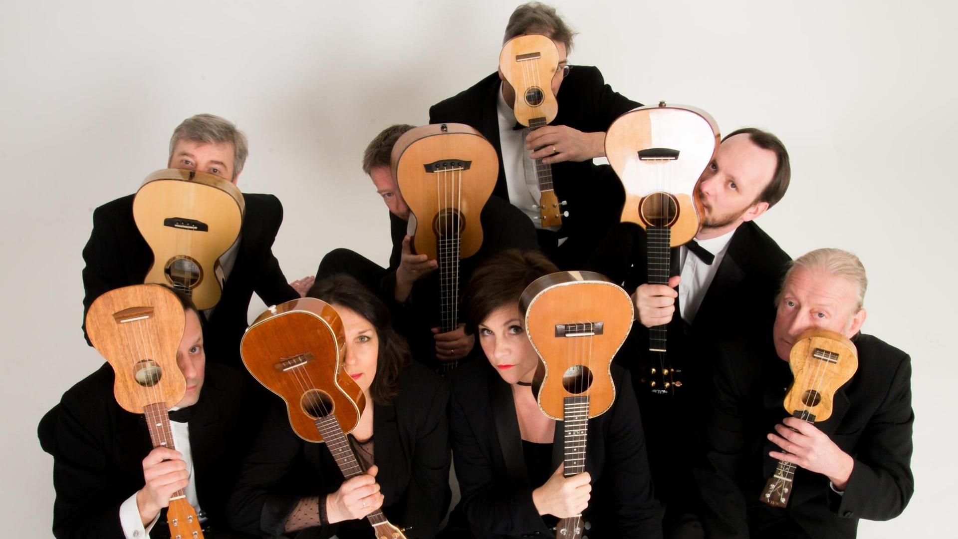 Die Mitglieder des Ukulele Orchestra of Great Britian stehend und sitzend und schwarz gekleidet verdecken mit ihren Instrumenten die Hälfte ihrer Gesichter.