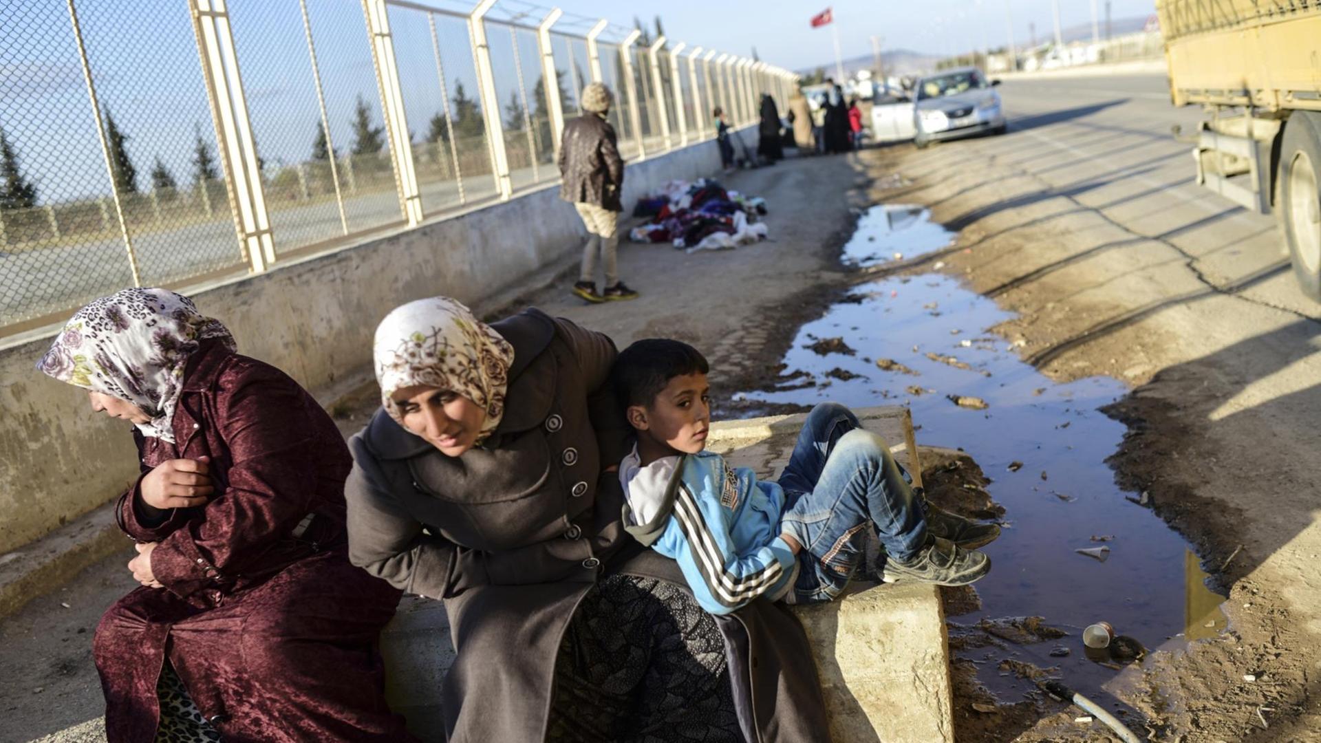 Zwei syrische Frauen und ein Junge warten in der Nähe des Tores von Öncüpınar vor einem Zaun.