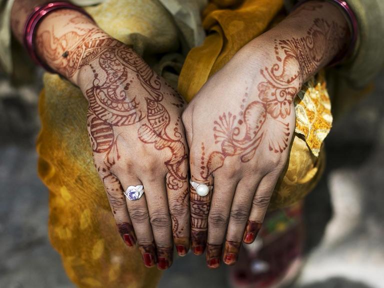Eine Frau zeigt ihre mit Henna bemalten Handoberflächen. Am linken Zeigefinger trägt sie einen Ring mit einem weißen Stein, am rechten Ringfinger einen Silberring mit lilafarbenem Stein.