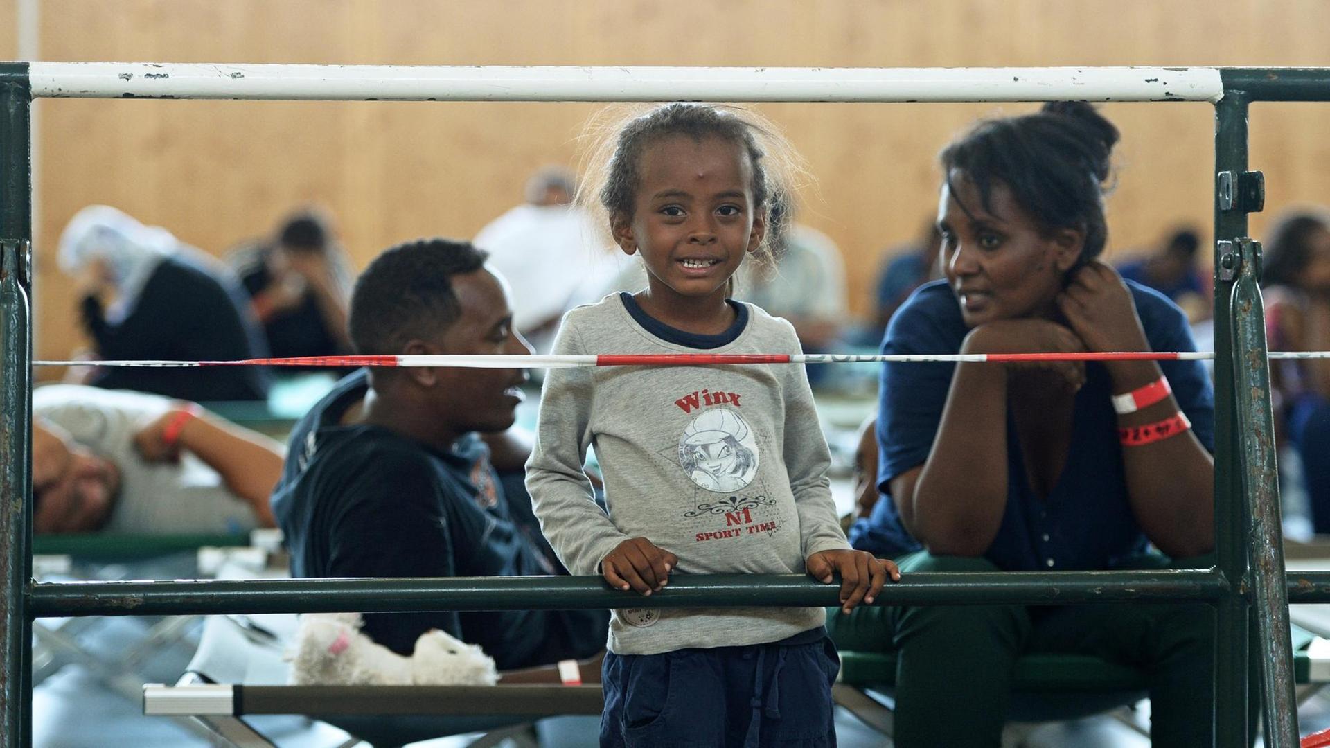 Ein Mädchen aus Eritrea steht neben seiner Mutter vor einem Absperrgitter in einer Turnhalle.