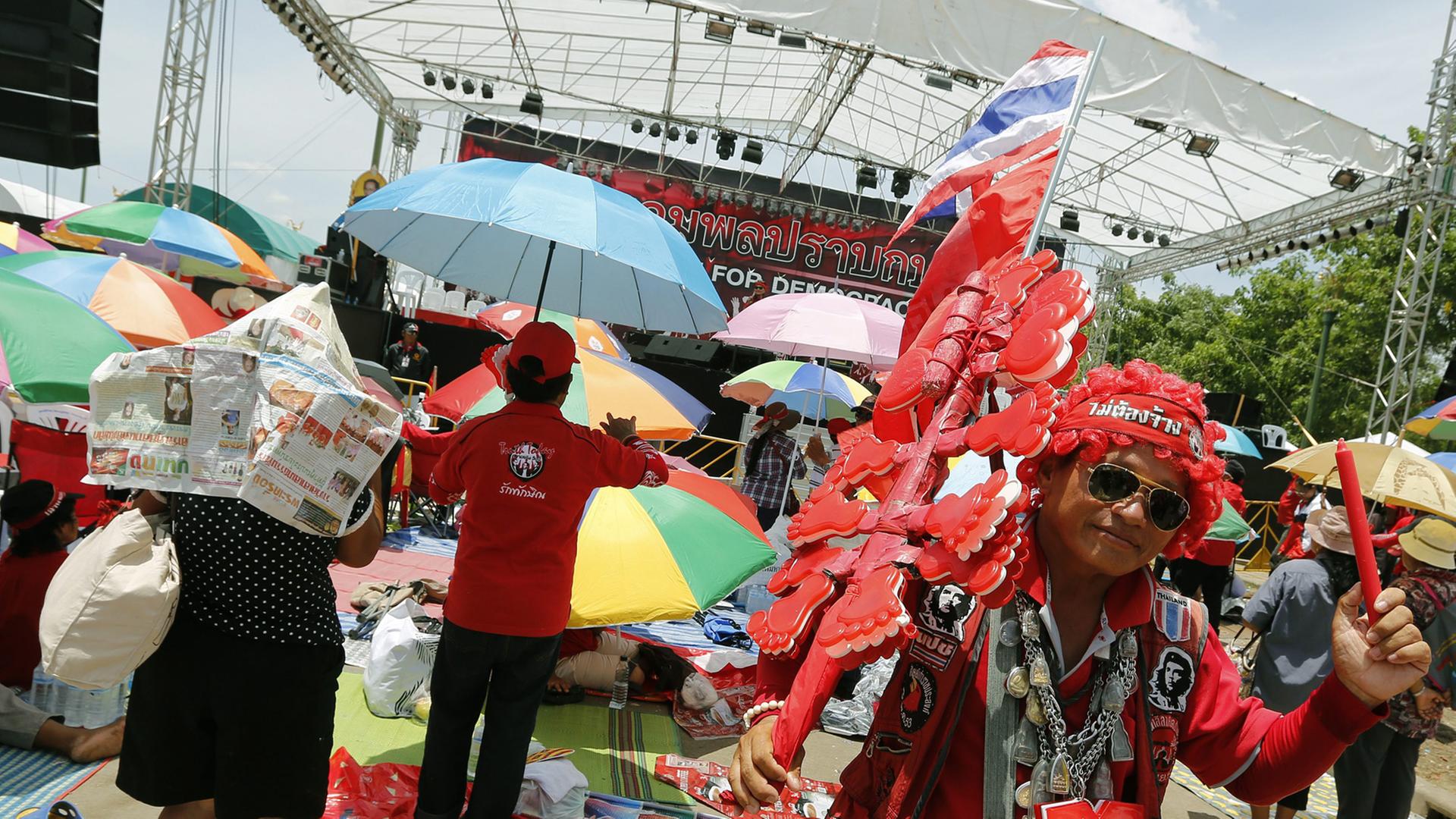 Ein in rot gekleideter Demonstrant mit Sonnenbrille und Thailand-Flagge schwenkt einen roten Stift, hinter ihm unter Sonnenschirmen Dutzende Menschen, die auf eine Bühne schauen