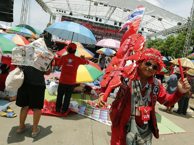 Ein in rot gekleideter Demonstrant mit Sonnenbrille und Thailand-Flagge schwenkt einen roten Stift, hinter ihm unter Sonnenschirmen Dutzende Menschen, die auf eine Bühne schauen