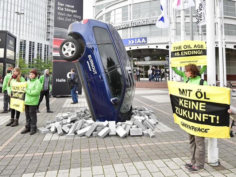 Aktivisten von Greenpeace neben einem Auto, das aussieht, als wäre es schräg in den Boden gerammt worden - als Protest gegen klimaschädliche Abgase.