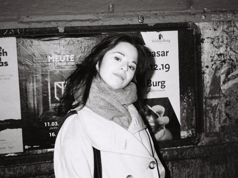 Schwarz-Weiß Aufnahme von Chiara Battaglia, lange dunkle Haare, heller Mantel, vor einer Wand mit Plakaten.