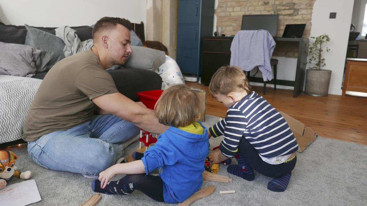 Ein junger Vater sitzt am Boden und spielt mit seinen zwei kleinen Kindern.