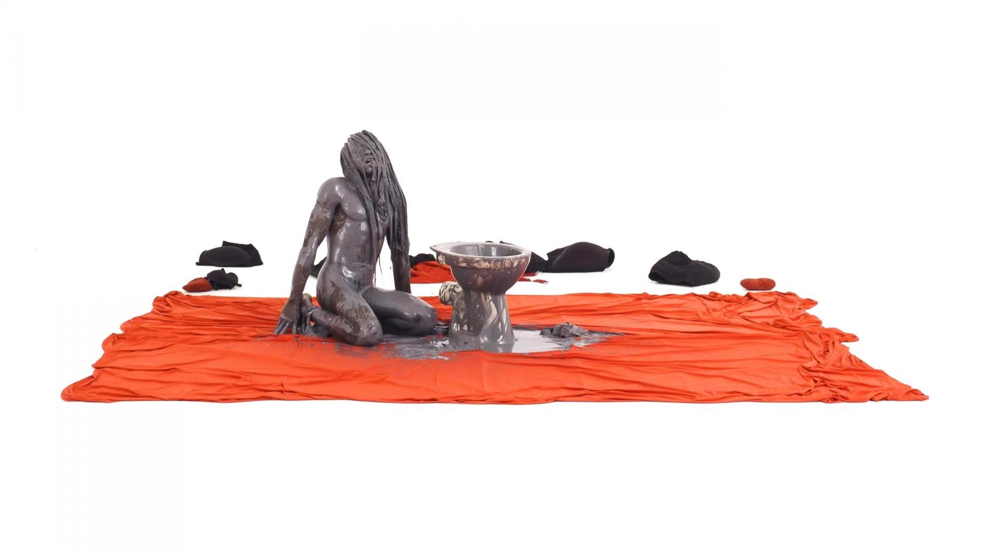 Die Performance "New Scenario" von Va-Bene Elikem Fiatsi: Eine Mensch in Marmor-Optik hockt auf einem roten Teppich vor einer Toilette - zu sehen in der VR-Ausstellung "Whiteout" im NRW-Forum Düsseldorf