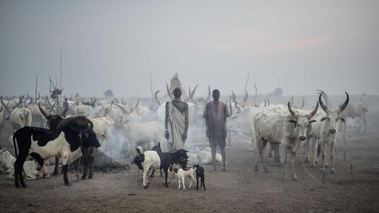 Zwei Mitglieder der Mundari aus dem Südsudan vor ihrer Rinderherde. Es ist dunstig, aus einer Feuerstelle steigt Rauch auf.