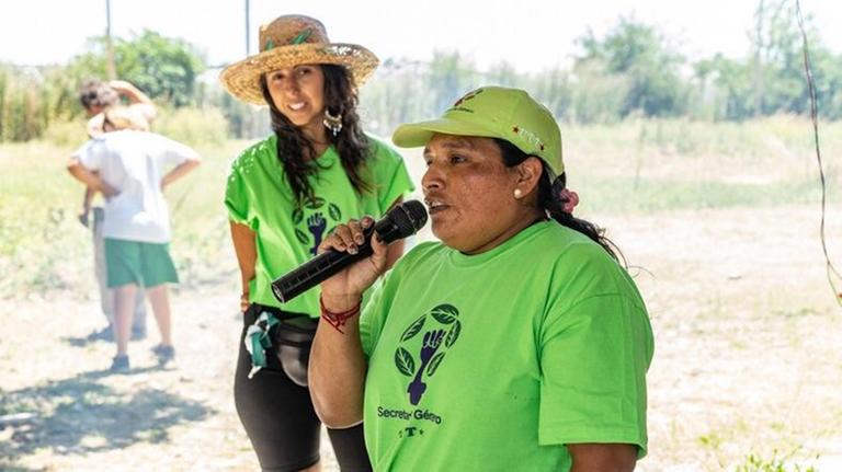 Eine ältere Frau mit Käppi, Zopf und knallgrünem T-Shirt spricht in ein Mikrofon, eine junge Frau mit Strohhut im Hintergrund hört zu.