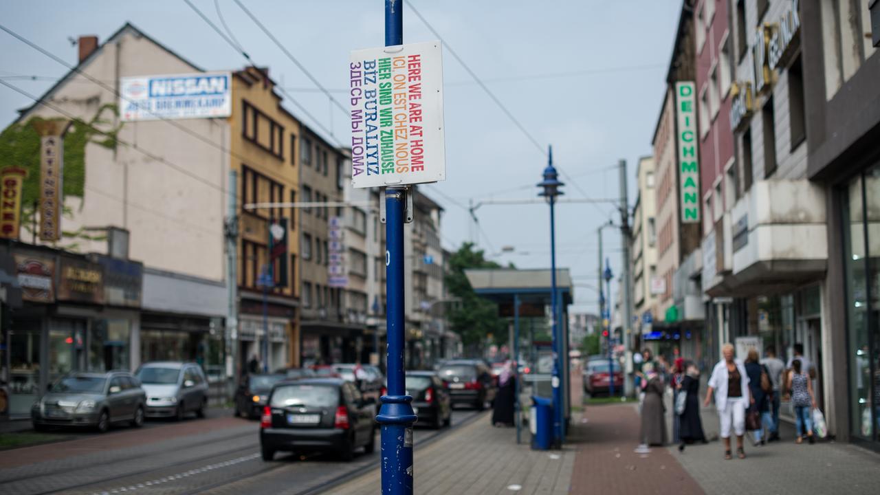 Ein Schild "Hier sind wir zuhause" in verschiedenen Sprachen hängt am 12.08.2015 in Duisburg (Nordrhein-Westfalen) auf der Straße. Händlern brechen die Geschäfte weg, Familienclans reklamieren die Gegend für sich.