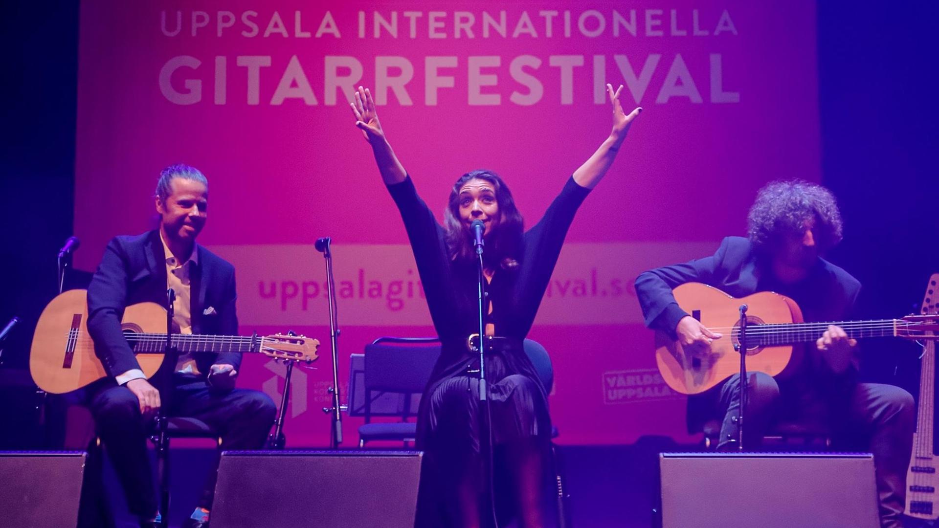 Drei Künstler sitzen auf der Bühne, die Sängerin in der Mitte, die beiden Gitarristen ihr zu Seite. Sie streckt beide Arme in die Höhe, im Hintergrund das Festivalplakat auf pinkfarbenem Grund: Uppsala Internationella Gitarrfestival