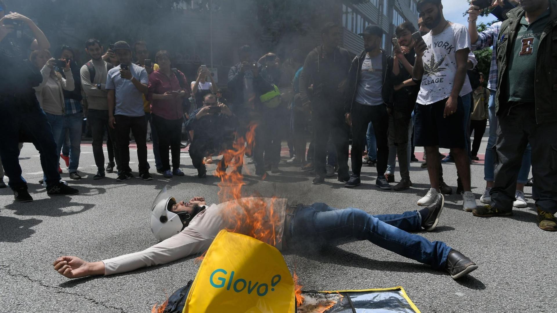 Ein Beschäftigter des spanischen Kurierdienstes Glovo liegt auf der Straße neben einem brennenden Glovo-Rucksack vor dem Gebäude des Unternehmens in Barcelona während einer Protestaktion am 27.05.2019 nach dem tödlichen Unfall eines Glovo-Fahrradkuriers.