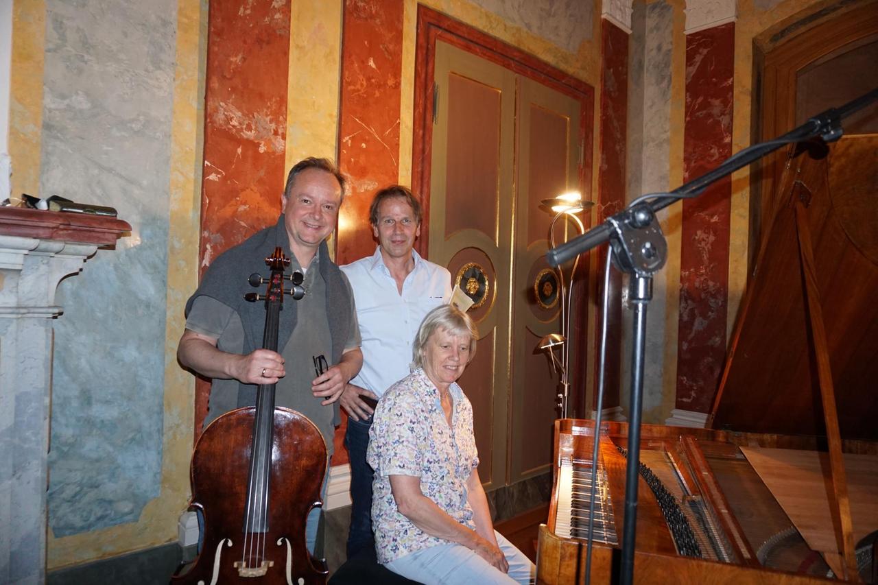 Der Cellist mit Cello in der Hand, der Moderator mit Noten unter dem Arm und  die Pianistin neben dem Hammerklavier auf einem Hocker schauen in die Kamera.
